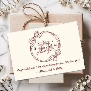 May include: A rectangular cream-colored greeting card featuring a dark red line-art illustration of two clinking champagne glasses inside a ribbon-tied circular frame. The text reads: Congratulations!! We are so happy for you! We love you! -- Allison, Bob & Bobby.
