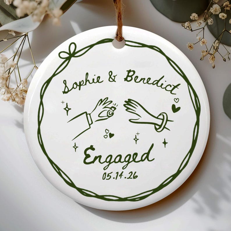 May include: A round white ceramic ornament featuring a dark green line-art wreath. Inside, the text reads Sophie & Benedict, Engaged, and 05.14.26. The design includes illustrations of two hands, one holding a ring, surrounded by small stars and hearts.