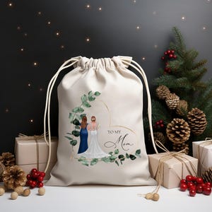 Puede incluir: Una bolsa de regalo beige con cordón y un diseño en forma de corazón con dos figuras y el texto "TO MY Mom". El diseño está enmarcado por hojas verdes. La bolsa está rodeada de regalos envueltos, piñas y un árbol de Navidad.