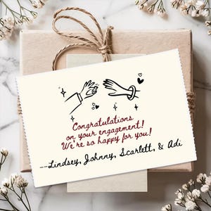 May include: A rectangular off-white greeting card featuring a black line drawing of two hands, one holding a ring, with the text: Congratulations on your engagement! We're so happy for you! --Lindsey, Johnny, Scarlett, & Adi, tied to a brown gift box.