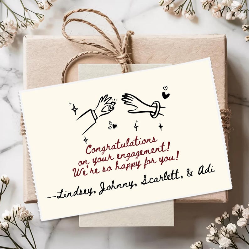 May include: A rectangular off-white greeting card featuring a black line drawing of two hands, one holding a ring, with the text: Congratulations on your engagement! We're so happy for you! --Lindsey, Johnny, Scarlett, & Adi, tied to a brown gift box.