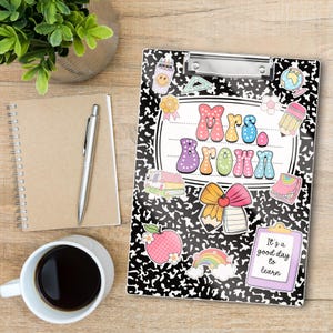 Op de afbeelding: Een zwart-wit klembord in compositieboekstijl met de naam "Mrs. Brown" in kleurrijke letters. Het klembord is versierd met diverse stickers met schoolthema's, waaronder een appel, een regenboog en een potlood.