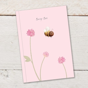 Puede incluir: Una tarjeta rosa con el texto "Busy Bee" escrito en cursiva negra. Una abeja de dibujos animados con rayas marrones y amarillas vuela sobre tres flores rosas con tallos verdes.