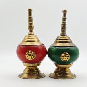 May include: Two decorative containers with brass bases and lids featuring a tall, ribbed finial. One container has a smooth, rounded red body, and the other has a smooth, rounded green body. Each features a central gold-toned floral emblem.