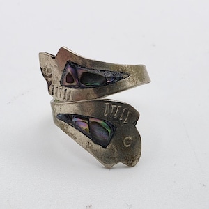 May include: A bypass style ring crafted from tarnished silver-toned metal, featuring two triangular inlays of iridescent abalone shell. The metal surface displays etched linear details and a weathered finish, with an open, adjustable band design.
