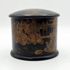 May include: A cylindrical black container with a lid, featuring gold-colored illustrations of figures in traditional attire, a fence, and architectural elements. The surface is decorated with detailed scenes of people sitting outdoors.