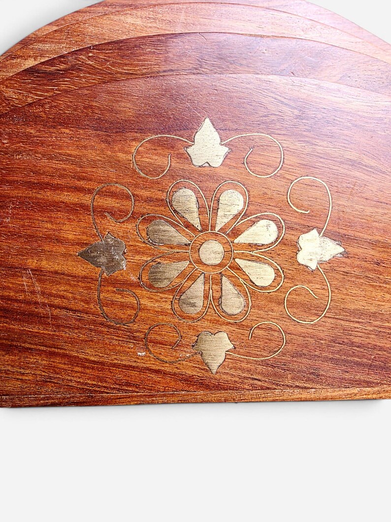 May include: A set of semi-circular wooden coasters featuring a rich brown grain and a central brass inlay. The inlay depicts a stylized flower with twelve petals surrounded by four leaf-shaped accents and thin, curved decorative lines.