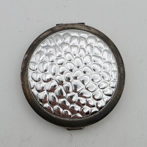 May include: A round compact mirror with a silver-toned, hammered, pebble-like texture on the lid. The outer frame of the compact is a dark, tarnished metal finish, showing a circular hinge at the top and a small latch at the bottom edge.
