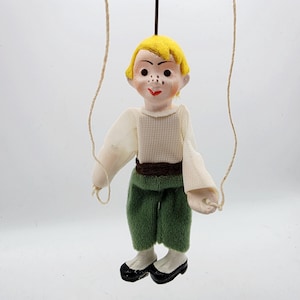 May include: A marionette puppet featuring a figure with bright yellow hair, a white long-sleeved shirt, a dark brown waistband, and green trousers. The puppet has painted facial features including freckles and red lips, and is suspended by strings.
