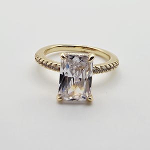 May include: A gold ring featuring a large, rectangular, clear faceted stone held by four prongs. The band is thin and metallic gold, accented with a row of small, clear, sparkling stones embedded along the sides of the shank.