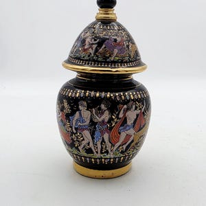 Può includere: Un vaso in ceramica nera con coperchio abbinato, decorato con figure classiche dipinte a mano nei toni del rosso, blu e bianco. Il contenitore presenta bordi dorati lungo l'apertura, la base e il coperchio, raffiguranti scene dettagliate di figure umane in un paesaggio.