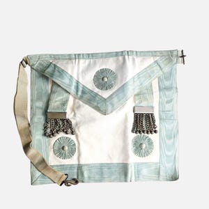 May include: A white ceremonial apron featuring light blue silk trim and a triangular flap. The apron is decorated with three circular pleated rosettes and two silver-toned metal tassels suspended from rectangular bars on the front surface.