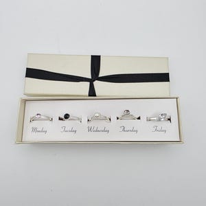 May include: A set of five silver-toned rings presented in a rectangular cream-colored box with a black ribbon. Each ring is labeled with a day of the week from Monday to Friday, featuring distinct stones including pink, black, pearl, and clear gems.