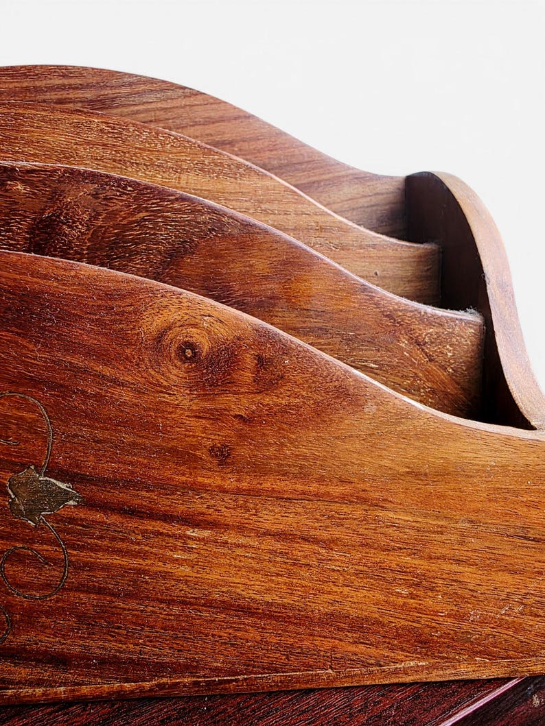 May include: A wooden letter holder featuring a tiered, wave-like design with multiple slots for organizing documents. The natural brown wood grain is visible throughout, and a small, metallic floral inlay is embedded on the front panel.