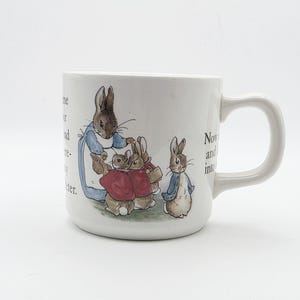 May include: A white ceramic mug featuring a colourful illustration of Peter Rabbit and other rabbits in blue jackets and red coats. The design includes black printed text excerpts from the story, a curved handle, and a smooth, cylindrical body.