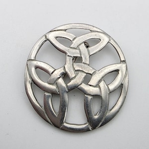 May include: A circular silver-toned brooch featuring an openwork design of an intricate, three-part interlaced knot pattern. The metal surface has a smooth, matte finish with visible contours that define the overlapping loops of the knotwork.