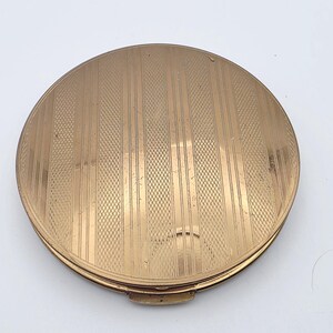 May include: A round, gold-toned metal compact case featuring a textured surface with alternating vertical stripes and a cross-hatched diamond pattern. The circular object has a smooth, polished edge and a small latch mechanism at the bottom.