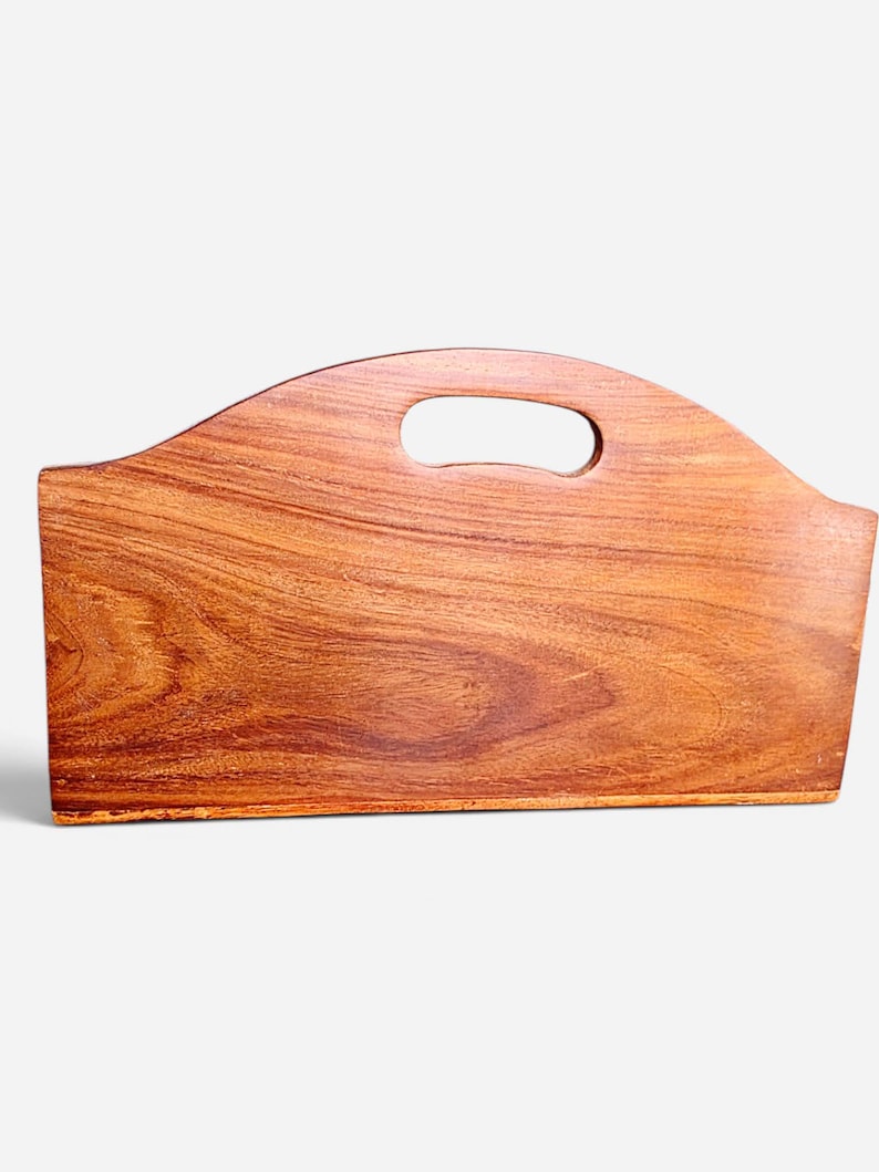 May include: A rectangular wooden magazine rack with a rich, warm brown grain pattern. The top edge features a smooth, arched design with a central oval cutout that serves as a handle for carrying. The structure is solid with a flat base.