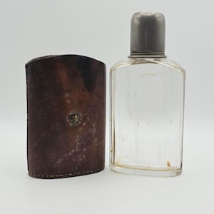 May include: A clear glass flask with a rounded metal cap positioned next to a dark brown leather case featuring a single metal stud closure. The flask has a rectangular body with rounded corners and a flat base, while the leather case is curved.