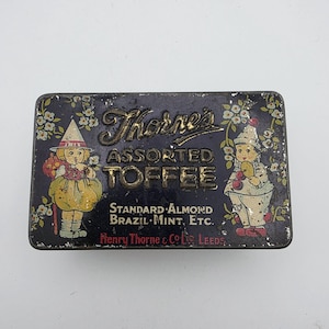 May include: A rectangular metal tin with a dark blue background featuring the text Thorne's Assorted Toffee, Standard Almond, Brazil, Mint, Etc., and Henry Thorne & Co. Ltd. Leeds. Two illustrated figures in pointed hats flank the central text.