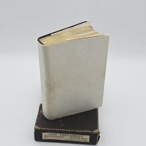 May include: A small book with a plain white dust jacket standing upright on top of its dark brown rectangular storage box. The box features a label that reads: NEW TESTAMENT PRONOUNCING MINION 48mo. Manufactured in Great Britain.