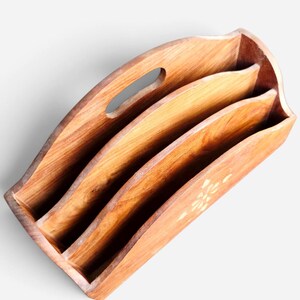 May include: A wooden magazine rack featuring a natural brown grain finish with three distinct storage compartments. The top edge has a curved design with an integrated oval handle cutout, and a small floral inlay detail is visible on the front.