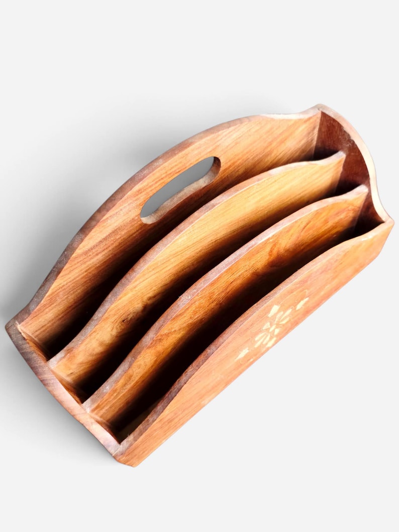 May include: A wooden magazine rack featuring a natural brown grain finish with three distinct storage compartments. The top edge has a curved design with an integrated oval handle cutout, and a small floral inlay detail is visible on the front.