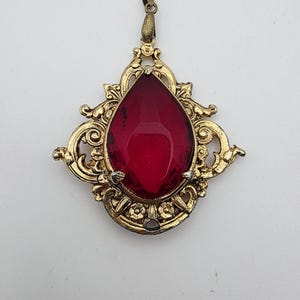 May include: A gold-toned metal pendant featuring an ornate, filigree-style frame with scrollwork and floral details. A large, faceted, pear-shaped deep red gemstone is set in the center, secured by small metal prongs at the base.