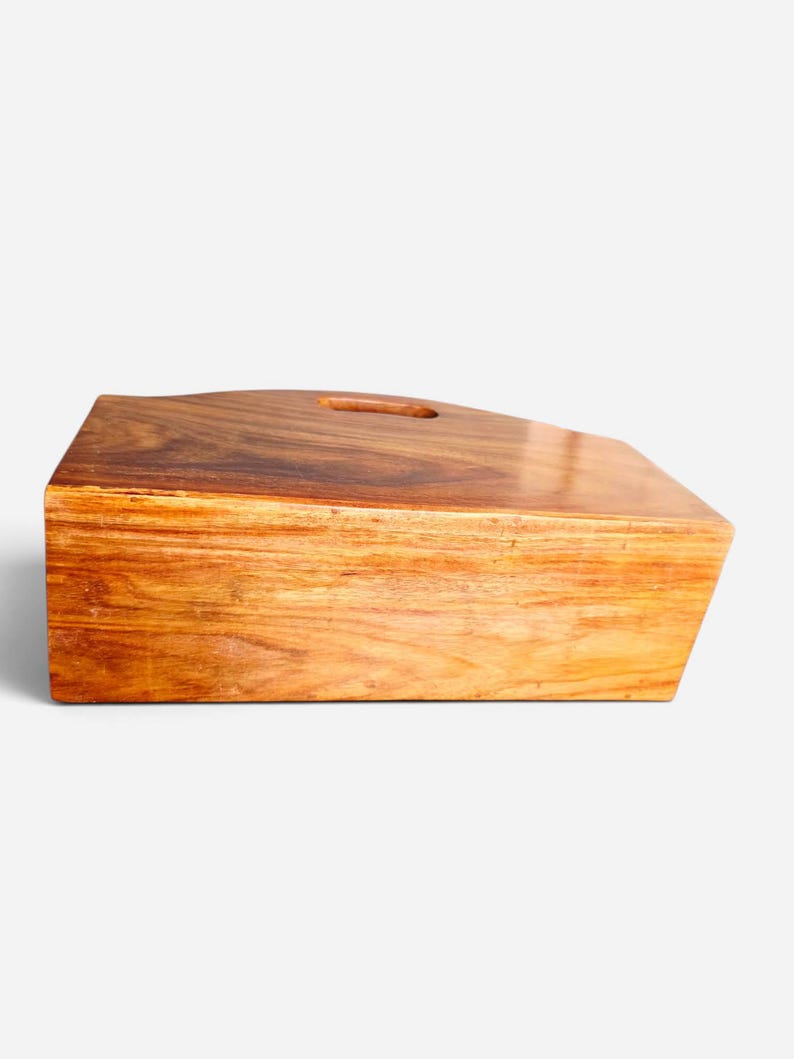 May include: A rectangular wooden serving board with a smooth, polished finish and a rich brown grain pattern. The board features a slightly angled top edge and a single oval-shaped cutout handle positioned near the center of the upper surface.
