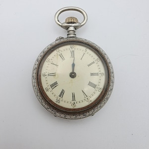 May include: A silver-toned pocket watch with an intricate floral engraved border and a cream-colored dial. The face features black Roman numerals, a minute track with Arabic numerals in increments of five, and two dark hands pointing to twelve.