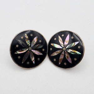 May include: A pair of circular earrings featuring a black background with an inlaid iridescent abalone shell flower design at the center. Each earring is accented with small silver-toned dots arranged in a circle around the floral motif.