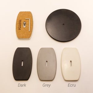 May include: Five different colored round and oval-shaped ceramic coasters. The coasters are in the colors dark grey, light grey, white, black, and cork. The coasters are labeled "Dark", "Grey", "Ecru", and "Cork".