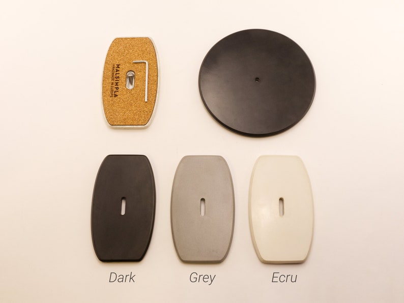May include: Five different colored round and oval-shaped ceramic coasters. The coasters are in the colors dark grey, light grey, white, black, and cork. The coasters are labeled "Dark", "Grey", "Ecru", and "Cork".