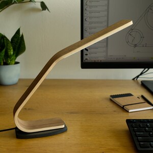 May include: A wooden desk lamp with a curved arm and a black base. The lamp is on a wooden desk with a computer monitor, keyboard, and notebook.