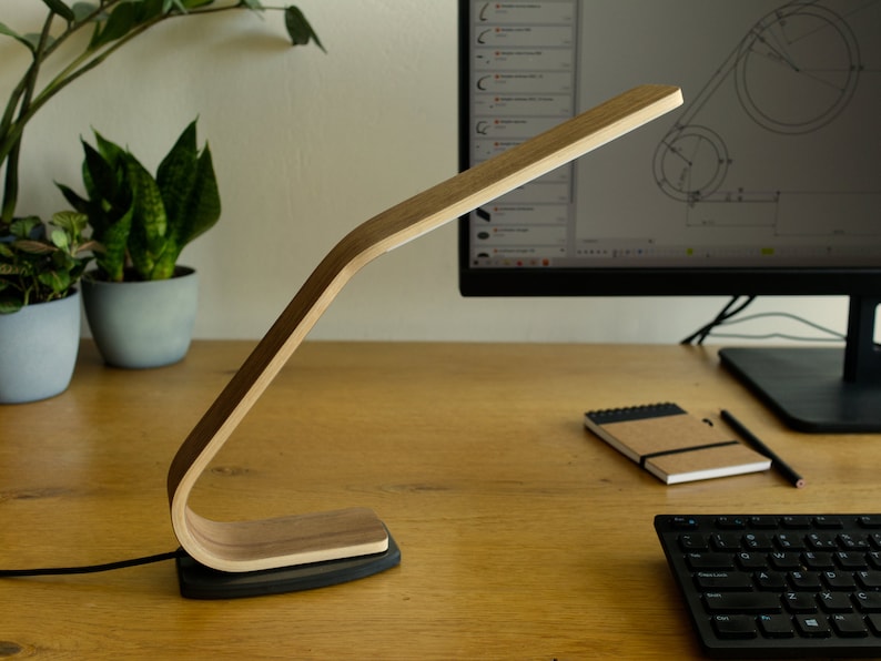 May include: A wooden desk lamp with a curved arm and a black base. The lamp is on a wooden desk with a computer monitor, keyboard, and notebook.
