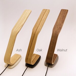 May include: Three wooden desk lamps in different wood finishes: ash, oak, and walnut. Each lamp has a curved wooden arm and a base with a cord.