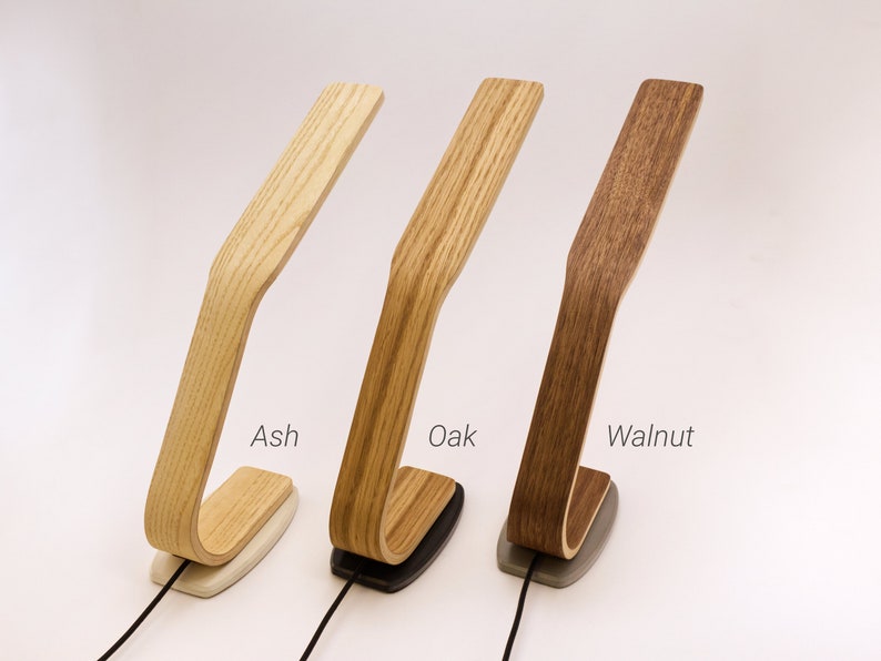 May include: Three wooden desk lamps in different wood finishes: ash, oak, and walnut. Each lamp has a curved wooden arm and a base with a cord.