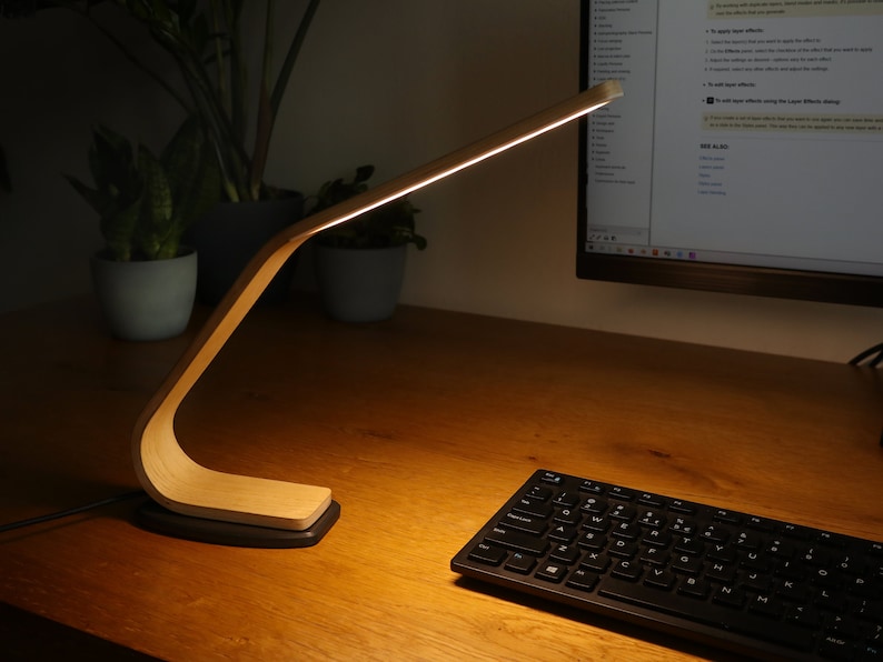 May include: A wooden desk lamp with a flexible arm and a warm white light. The lamp is on a wooden desk with a keyboard and a potted plant.