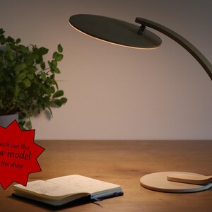 May include: A wooden desk lamp with a circular light shade. The lamp has a curved arm and a round base. The lamp is on a wooden table with a notebook and a plant in a pot.
