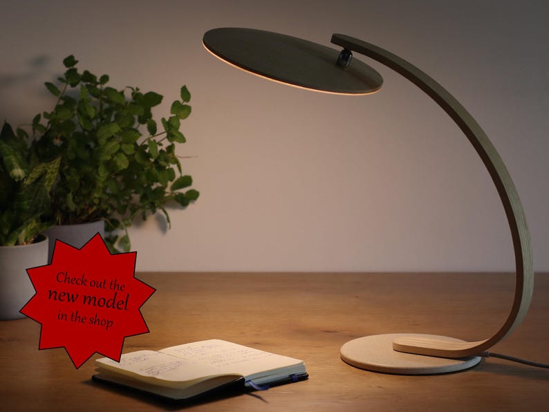 May include: A wooden desk lamp with a circular light shade. The lamp has a curved arm and a round base. The lamp is on a wooden table with a notebook and a plant in a pot.