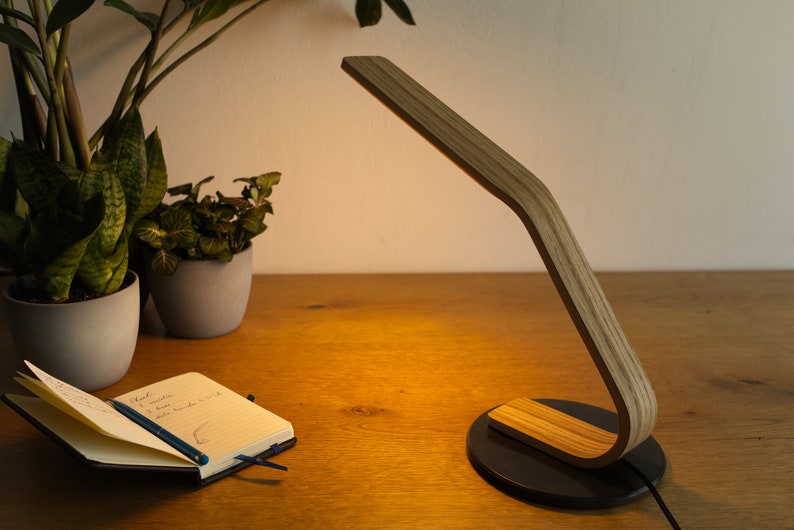 May include: A wooden desk lamp with a curved arm and a black base. The lamp is on a wooden desk with a notebook and a pencil on it. There are two potted plants in the background.
