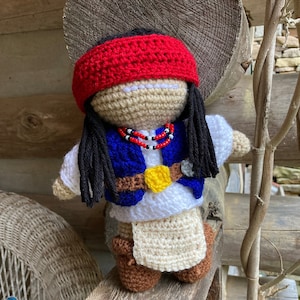 May include: Crocheted doll with long black hair, wearing a red headband, a white shirt, a blue vest with a yellow star, and brown boots. The doll is holding a beaded necklace.