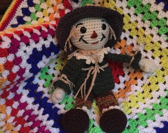 May include: A crocheted scarecrow doll with a brown hat, green shirt, and brown shoes. The doll is sitting on a colorful crocheted blanket.