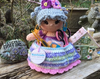 May include: A crocheted doll with gray hair and a pink dress is holding a container of sewing supplies. The doll is sitting on a wooden surface with a small stone garden gnome and a small house figurine. The doll's skirt has a tag that reads "Handmade with care by Sandy Northwall Eggert".
