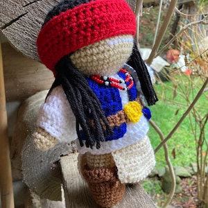 May include: Crocheted doll with long black hair, wearing a red headband, a white shirt, a blue vest, a brown belt with a yellow buckle, and brown boots. The doll is holding a beaded necklace.