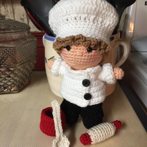 May include: A crocheted doll dressed as a chef, wearing a white chef's hat, white shirt, and black pants. The doll is holding a red and white rolling pin and a white spoon. The doll is sitting on a red and white baking mat with the words "Made in France / Roll'P" printed on it.