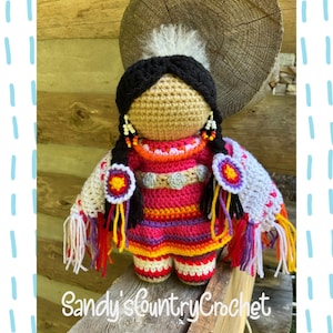 May include: A handmade crochet doll featuring a tan face, black yarn hair in braids with white and orange beads, and a vibrant pink dress with multi-colored stripes. The doll wears a white shawl with fringe and the text Sandy'sCountryCrochet.