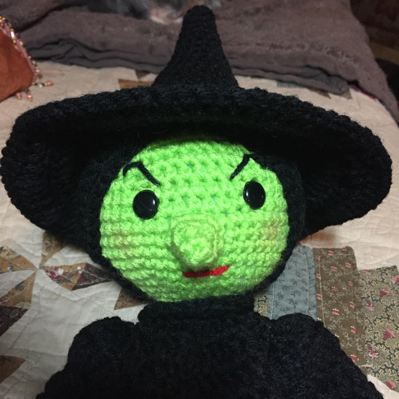 May include: A green crocheted witch doll with a black pointed hat and a large, crooked nose. The doll has black eyes and a red mouth.