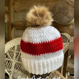 Op de afbeelding: Witte gehaakte muts met een rode streep en een bruine nep bont pompon. De muts ligt op een houten oppervlak. "SandysCountryCrochet" staat op het oppervlak.