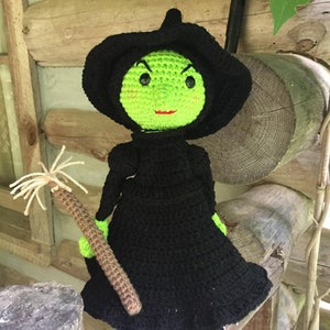 May include: A green crocheted witch doll with black yarn hair and a black hat. The doll is holding a brown crocheted broom.